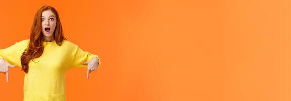 Take look quick. Amazed and fascinated cute ginger girl, redhead student in yellow sweater, gasping open mouth from surprise and wonder, pointing fingers down, look impressed, orange background photo