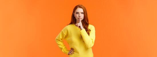 Serious and mad elder sister say keep silent, forbid talking during class, shushing angry with index finger pressed mouth, hold hand on waist in dissatisfied pose, standing orange background photo