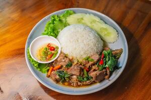 Stir-fried beef with basil in a blue plate with fish sauce, chili, cucumber and lettuce on a wooden table. photo
