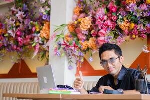 Asian male businessman to camera using laptop computer to work on project assignment from client hand thumbs up okay sign at office desk, sitting at workplace cafe photo