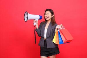chinese sales woman shouts cheerfully candidly through a megaphone speaker and lifts a shopping paper bag wearing a gray jacket on a red background. for transaction, lifestyle and business concepts photo
