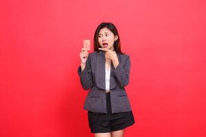 Asian office woman's expression shocked candid hand holding a debit credit card while pointing at it wearing a jacket and skirt on a red background. for financial, business and advertising concepts photo