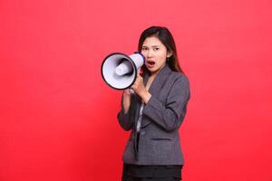 Asian office girl expression is very angry screaming using a megaphone in front of her wearing a gray jacket and skirt on a red background. for broadcast, business and advertising concepts photo