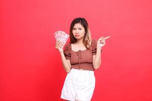 The expression of a sullen Asian girl holding rupiah money while pointing to the left wearing a brown blouse with a red background. for transaction, technology and advertising concepts photo