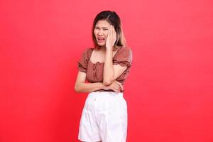 The expression of an Asian woman with her arms around her head is very painful and dizzy looking at the camera wearing a brown blouse top and shorts with a red background. for health and advertising photo