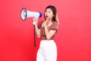 the expression of an indonesia woman screaming in surprise at the camera holding an audio megaphone while focusing on the sound wearing a brown blouse top and shorts on a red background technology photo