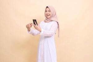 Asian Muslim woman wearing a hijab smiling happily winking at the camera holding a cell phone gadget in her hand while pointing at it. Lifestyle, technology and promotion concept photo