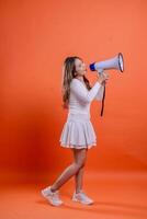 full length indonesia ladies white top standing left candid shouting cheerful both hands holding megaphone loudspeaker isolation orange background. for technology, advertising, broadcast photo