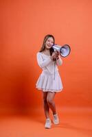 full length beautiful asian girl white top standing to camera screaming cheerful both hands holding megaphone loudspeaker isolated orange background. for technology, advertising, broadcast, promotion photo