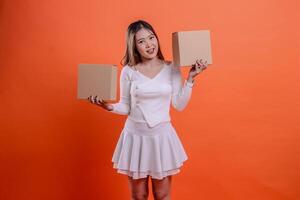 Beautiful adult female in white top happy to camera both hands lifting plain cardboard box up and down, standing on orange background. for delivery concept, gift, expedition, service, package photo