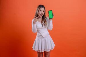 expression of beautiful teenage girl in white top standing cheerfully to camera hand holding mobile phone screen, cellphone while pointing at it isolated orange background. for technology, business photo