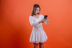 gesture beautiful young asian woman white top standing surprised excited to gadget slanted left both hands hold mobile phone, cellphone isolated orange background. for communication concept, business photo