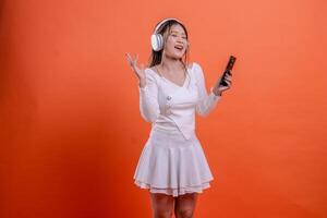 woman white top singing close eyes using earphone headphone while holding mobile phone hand sloping upward, cellphone, standing on orange background. for entertainment, lifestyle, business, service photo