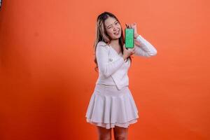 gesture young indonesia woman in white top standing cheerful winking to camera both hands holding mobile phone screen, cellphone isolated orange background. for technology, business, promotion photo