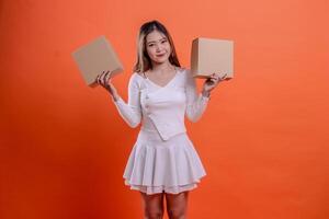 Beautiful young Asian girl in white top smiling to camera with both hands holding plain cardboard box, standing on orange background. for shipping concept, gift, expedition, service photo