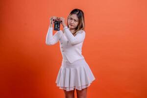charm adult beautiful woman white top standing pouting both hands holding car key keyless system isolated orange background. for transportation, rental services, vehicle buying and selling photo