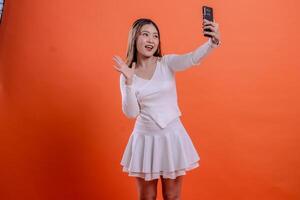 cheerful adult asian woman in white sweater call using smartphone camera, mobile phone, cellphone while pointing at face, standing. for technology, lifestyle, business, service photo