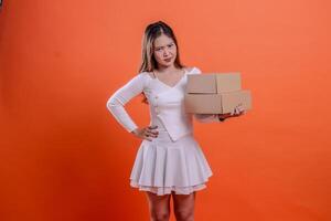 portrait of young asian woman in white top angry at camera hands carrying 2 plain cardboard boxes and holding waist, standing on orange background. for delivery, gift, expedition, service, package photo