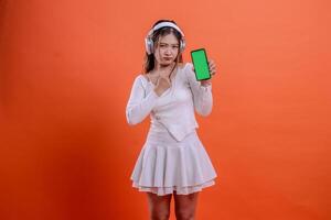 adult female in white sweater pouting wearing earphone headphone, holding mobile phone screen, cellphone green screen while clenching, standing. for entertainment, lifestyle, business, service concept photo