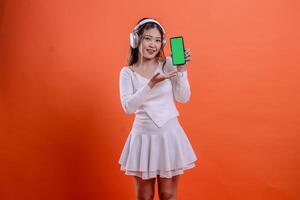 cheerful woman in white sweater wearing earphone headphone while holding mobile phone screen, cellphone green screen while presenting, standing on orange background. for entertainment, service concept photo