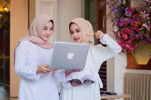 a pair of young female wearing hijab smile looking at the notebook screen while pointing holding the laptop and carrying a book standing in the outdoor area of restaurant. for technology, lifestyle photo