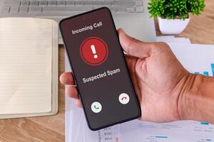 Hand holding a smartphone with incoming call from unknown caller on the screen at workplace. Spammer incoming call concept. photo