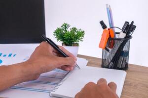 A man writes with his left hand in a blank notebook at a work desk with laptop, stationery, paperwork and plants in pot photo