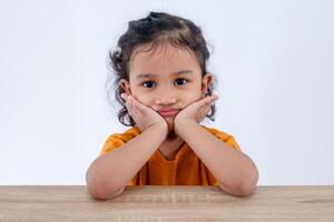 A little Asian girl with a pouting face sitting at table and posing with her hand on chin photo