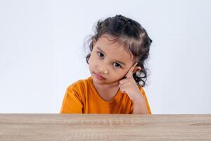 Adorable little Asian girl sitting at table and posing with one hand pointing to her head. Confused, thinking, or dreaming concept. photo