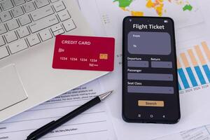 Office desk with credit card, smartphone with online airline ticket booking application on screen, laptop, paperwork, and pen photo
