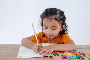 A smart little Asian girl learning to write in a notebook with wooden alphabet letter puzzles on a table photo