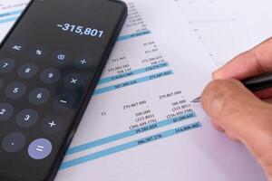 Close up of hand holding a pen and calculating financial data using calculator app on mobile phone. An accountant analyzing financial report data. photo