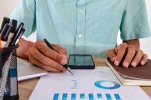 Man holding a pen and analyzing a graph of a report at workplace with laptop, notebook, documents, and mobile phone on the table photo