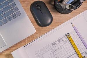 Top view of an architect's work desk with building layouts, stationery, pencil, laptop, mouse, and roll up metal tape measure photo