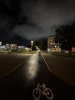The image shows a night scene of an urban area with a paved road leading towards buildings in the distance. There is a bicycle lane symbol painted on the road, and street lights illuminate the scene. photo