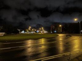 The image shows a nighttime cityscape with a wet road reflecting street lights. There are buildings in the distance, street lamps along the road, and a cloudy sky above. photo