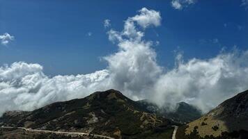 A captivating view of clouds rolling over a lush mountain range, with a winding road cutting through the green hills, under a bright blue sky. photo