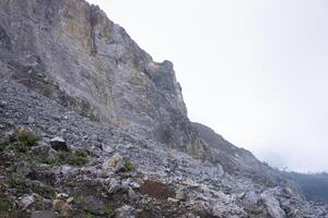 A rugged rocky mountainside under a cloudy sky, showing steep, barren cliffs with loose stones scattered across the terrain. Minimal vegetation clings to the inhospitable environment. photo