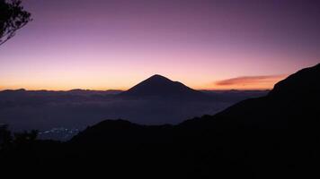 A silhouetted mountain peak stands against a gradient sky transitioning from deep purple to soft orange at twilight. The dark foreground hills and distant lights from a valley add depth to the serene photo