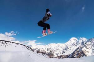 Female snowboarder jumping off a ski jump and doing a trick. Amazing shot of a snowboarder flying in the air and grabbing a trick against the backdrop of snowy mountains photo
