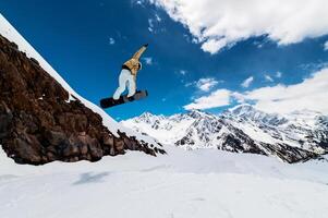 Professional snowboarder in helmet and ski clothes doing a trick in a jump against the background of high snowy mountains. Wide angle. Sports shooting photo