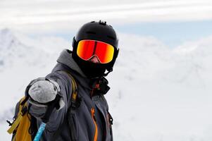 man points his hand at the camera in sunglasses with a ski helmet on his head and holds a stick in his hand against the background of white snow and mountains photo