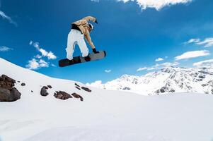 snowboarder jumps from a big air with a board grab, a nose or tail trick in rotation, against the backdrop of snow-capped mountains and the bright sun photo