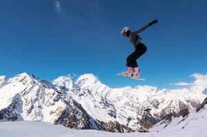 snowboarder jumps from a big air with a board grab, a nose or tail trick in rotation, against the backdrop of snow-capped mountains and the bright sun photo
