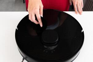 Robot vacuum cleaner with round black body on white background table, new home assistant, woman hand reaches out to turn it on and check, close-up photo