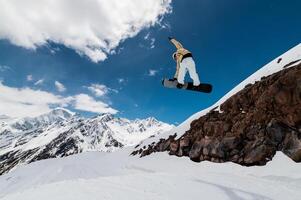 Snowboarding in high mountains. Male athlete doing a trick while jumping from a ski jump against the backdrop of mountains and a sunny day, spectacular shot photo
