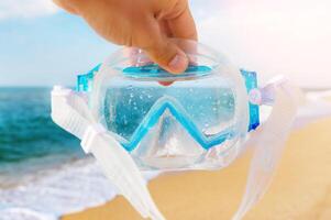 A man holds a snorkel mask in his hand against a clear blue sky and turquoise ocean. Diving mask in a man's hand. Photo of a mask and snorkel for swimming in a pool or sea with a central composition