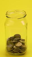 A clear glass jar filled with coins on a bright yellow background symbolizes savings and financial planning. This colorful setting emphasizes the importance of saving money. video