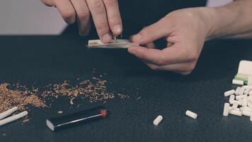 Man in Black Clothing is Rolling a Hand Made Cigarette Using Papers, Filters and Tobacco. A Way to Save on Tobacco Products. Tobacco Product Economy Concept - Slow Motion. Close-Up Shot video