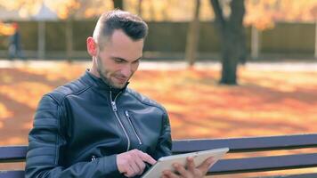 Student is Reading News or Watching on Tablet Computer in the Park in Autumn. Man in Black Leather Jacket is Sitting on a Bench Using Big White Tablet PC. Middle Close-Up Shot video